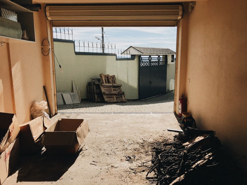 The image depicts a cluttered indoor space resembling a workshop or storage area, with a narrow pathway strewn with various discarded items such as cardboard boxes, plastic bags, and broken equipment.