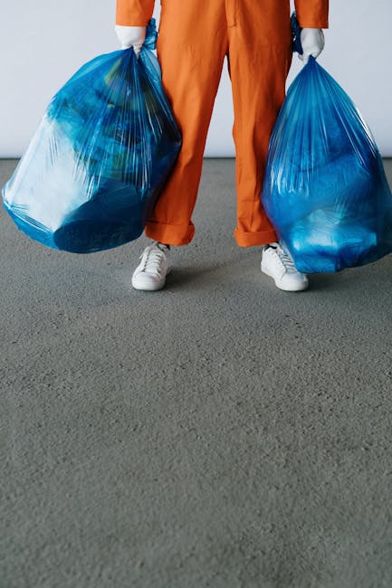 A person wearing a grey and yellow checked shirt, grey trousers, and bright green gloves stands outdoors on a grassy area, holding open a large black garbage bag made of plastic. The bag appears to be