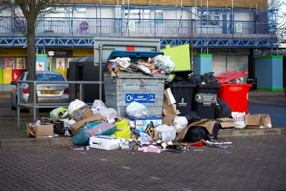 A large assortment of overflowing rubbish bins and scattered waste bags, cardboard boxes, paper, plastic packaging, and discarded household items are piled on a paved parking area. The debris includes