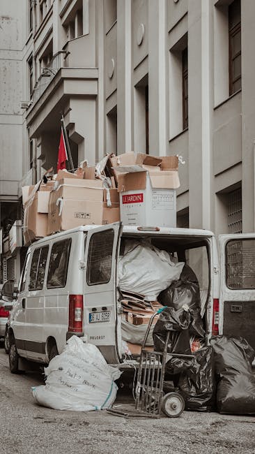 A large pile of flattened cardboard boxes and packaging materials stacked high behind a metal railing on a city street. The cardboard varies in shades of brown and is held together with plastic straps